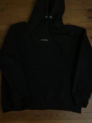 Svart hoodie från HOMME - Svart hoodie från HOMME med minimalistisk vit text på bröstet. Klassisk modell med huva och dragsnören samt magficka framtill. Perfekt för en clean och stilren look. Mjuk och skön bomullsblandning.