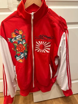 Röd Ed Hardy ziptröja med tryck - Jackan på bilden är en vintage Ed Hardy by Christian Audigier "Japan Edition" träningsjacka. Den kännetecknas av sin klarröda färg med vita ärmar som har kontrasterande röda ränder. Storlek. Storlek M för herrar.