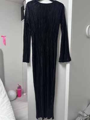 Svart långklänning i plisserat tyg - Snygg svart långklänning med plisserat tyg och långa ärmar. Klänningen har en rak passform och rund halsringning, perfekt för dig som gillar en stilren och elegant look.