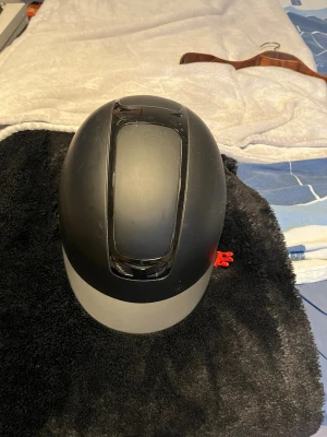 Svart ridhjälm från Horka - Säljer en svart ridhjälm från JH Collection med matt finish och ventiler upptill. Hjälmen har justerbart hakband i svart konstläder och mjuk vaddering på insidan för extra komfort. Passar perfekt för ridning och har en klassisk, rundad form.