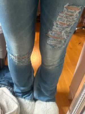 Blå slitna bootcut jeans - Säljer ett par blå bootcut jeans med kraftiga slitningar och hål på låren och knäna. Jeansen har en klassisk femficksmodell och ljusa tvättade partier framtill. Perfekta för en avslappnad och trendig look.