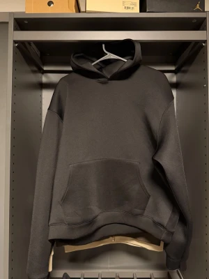 Svart basic hoodie med magficka - En stilren svart hoodie med klassisk huva och stor magficka framtill. Hoodien har en avslappnad passform och ribbade muddar vid ärmar och midja. Perfekt för en chill och enkel look. Tillverkad i mjukt material för extra komfort (scuba) material.