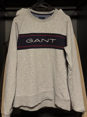Grå sweatshirt från GANT - Säljer en stilren grå sweatshirt från GANT med ett brett mörkblått band och röd detalj över bröstet där loggan är tryckt i vitt. Tröjan har rund halsringning, långa ärmar och ribbade muddar. Perfekt för en avslappnad och snygg look.