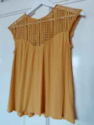 Gul ärmlös blus med spetsdetaljer - Snygg orange blus med ärmlös design och vackra spetsdetaljer upptill. Toppen har ett luftigt fall och är tillverkad i ett lätt, strukturerat tyg som passar perfekt till varma dagar. Den har rund halsringning och ett bohemiskt uttryck.