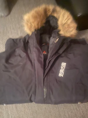 Svart dunjacka 8848 Altitude - Snygg svart dunjacka från 8848 Altitude med avtagbar huva och fuskpäls. Jackan har dragkedja framtill, hög krage och logga på bröstet. Perfekt för kalla vinterdagar tack vare det tjocka fodret och den klassiska designen.