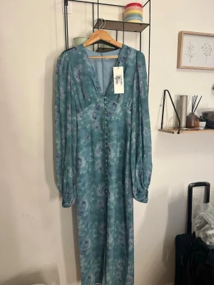 Blommig långklänning från Adoore - Långklänning i ljusblått med blommigt mönster från Adoore . Klänningen har v-ringning, knappar framtill och långa ärmar med lätt puff. Tillverkad i ett tunt, flowy material som ger en romantisk känsla. Dusty blue