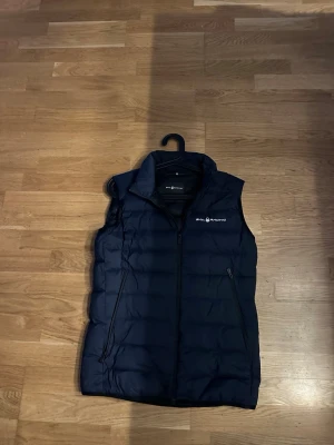 Mörkblå dunväst från Sail Racing - Snygg mörkblå dunväst från Sail Racing med hög krage och dragkedja framtill. Västen har två sidofickor med dragkedja och logga på bröstet. Perfekt för lager-på-lager och håller dig varm under kyliga dagar.