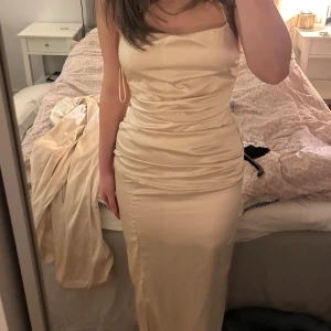 Champagnefärgad långklänning i satin - Elegant långklänning i champagnefärgad satin med figurnära passform och smala axelband. Klänningen har en mjukt draperad överdel och slät kjol som faller snyggt längs kroppen. Perfekt för speciella tillfällen.