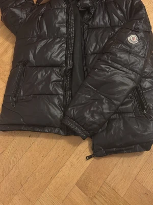Svart dunjacka från Moncler - Snygg svart dunjacka från Moncler med glansig finish och klassisk Moncler-logga på ärmen. Jackan har dragkedja framtill, två sidofickor med dragkedja och en huva. Perfekt för kalla vinterdagar och riktigt stilren look.