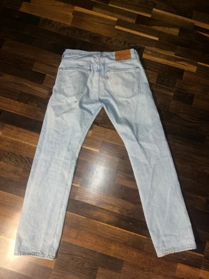 Ljusblå Levi's 501 med slitning - Säljer ett par ljusblå raka jeans från Levi's i klassisk modell. Jeansen har en tydlig slitning och hål på vänster lår, fem fickor och läderpatch bak i midjan. Tillverkade i bomullsdenim och har den ikoniska röda Levi's-lappen på bakfickan.