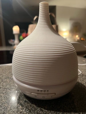 Vit aromadiffuser med sladd - Säljer en stilren vit aromadiffuser i plast med räfflad yta och droppformad design. Den har flera timerinställningar och knappar för ljus och av/på. Perfekt för att sprida doft eller fukt i rummet. Strömkabel medföljer.