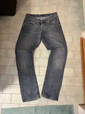 Blå raka jeans från Nudie Jeans - Säljer ett par klassiska blå raka jeans från Nudie Jeans med fem fickor och snygga slitningar. Jeansen har kontrastsömmar och broderade bakfickor. Tillverkade i slitstark denim, perfekta för en avslappnad streetstil.