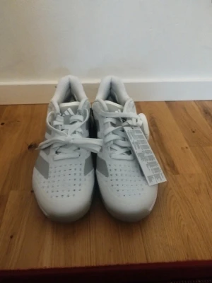 Vita Adidas sneakers med grå detaljer - Säljer ett par vita Adidas sneakers med grå ränder på sidorna och ventilerande hål i ovandelen. Skorna har klassisk snörning och en sportig design med stabil hälkappa. Perfekta för dig som gillar stilrena och bekväma sneakers.