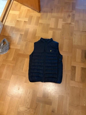 Svart dunväst från Lyle & Scott - Snygg svart dunväst från Lyle & Scott med gul logga på bröstet. Västen har ståkrage, dragkedja framtill och quiltad design. Perfekt att slänga över en hoodie eller tröja för extra värme och stil.