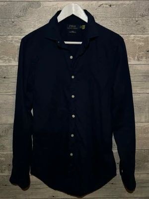 Mörkblå skjorta från Polo Ralph Lauren - Klassisk mörkblå skjorta från Polo Ralph Lauren med knappar framtill och lång ärm. Skjortan har en stilren design och är tillverkad i bomull, perfekt för en clean och tidlös look.