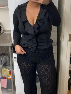 Svart topp med volang och v-ring  - Trendig svart omlottopp med långa ärmar och volangdetaljer längs urringningen och framtill. Toppens figurnära passform och djupa V-ringning ger en snygg siluett. Perfekt att styla med höga byxor eller kjol.