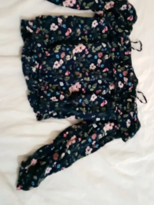 Blommig blus från H&M med puffärm - Mörkblå blus från H&M med blommigt mönster i rosa, vitt och grönt. Blusen har långa puffärmar och resår vid ärmslut och midja. Tillverkad i ett lätt och luftigt material, perfekt för en trendig och avslappnad look.