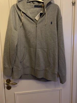 Ralph Lauren kofta - Klassisk grå hoodie med dragkedja från Ralph Lauren. Hoodien har två fickor fram, justerbar huva med vita snören och den ikoniska blå loggan broderad på bröstet. Tillverkad i mjukt sweatshirtmaterial, perfekt för chill dagar.