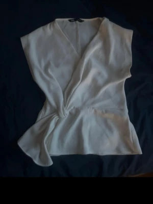 Vit omlottblus från Zara - Stilren vit omlottblus från Zara med djup V-ringning och knutdetalj i sidan. Blusen är ärmlös och har en luftig, lätt tygkänsla som ger en avslappnad men chic look. Perfekt att matcha med jeans eller kjol för en trendig outfit.