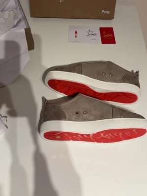 Gråa sneakers från Christian Louboutin - Snygga gråa sneakers i mocka från Christian Louboutin med den ikoniska röda sulan och vita kanter. Lågt skurna och minimalistisk design som passar till det mesta. Kommer med originalkartong och dustbags.