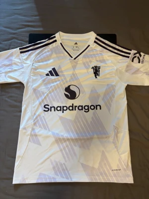 Manchester United bortatröja Adidas - Snygg vit Manchester United bortatröja från Adidas med lila detaljer och tryck. Tröjan har V-ringning, klubbmärke på bröstet, Snapdragon-logga och nummer 8 med B. Fernandes på ryggen. Tillverkad i lätt och ventilerande material med Aeroready-teknologi.