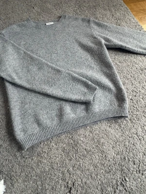 Grå stickad tröja med rund hals - En enkel och stilren grå stickad tröja med rund hals och långa ärmar. Tröjan har ribbstickade muddar vid ärmslut och nederkant. Perfekt för lager-på-lager och passar till jeans eller chinos. Materialet är mjukt och bekvämt.