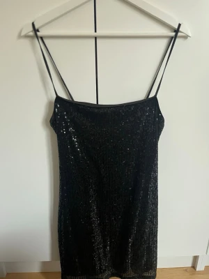 Svart paljettklänning med smala band - En snygg svart kortklänning med paljetter och smala axelband. Klänningen har en rak form och är perfekt för dig som vill sticka ut med lite extra glitter. Passar dig som gillar att klä dig trendigt och våga ta plats.