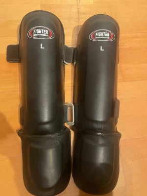 Svarta benskydd Fighter L - Svarta benskydd från Fighter Gear Genuine i storlek L, perfekta för kampsport eller träning. Tillverkade i slitstarkt syntetmaterial med vaddering för extra skydd. Justerbara kardborreband för bra passform och enkel användning. Aldrig använda är som ett par helt nya!