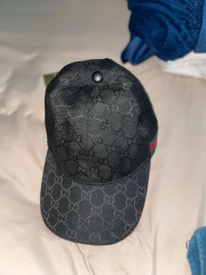 Svart Gucci keps med GG-mönster - Snygg svart keps från Gucci med klassiskt GG-monogram över hela kepsen. På sidan finns den ikoniska gröna och röda randen. Kepsen har böjd skärm och är tillverkad i ett slitstarkt tygmaterial. Perfekt för dig som vill ha en exklusiv och trendig accessoar.
