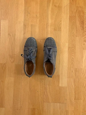 Grå mocka sneakers med nitar - Säljer ett par grå sneakers från Christian Louboutin i mocka med coola nitar på tån och klassisk röd sula. Skorna har snörning och en rund tå, vilket ger en edgy men stilren vibe. Perfekta för dig som vill sticka ut med exklusiva detaljer och lyxig känsla.Passar storlek 44.