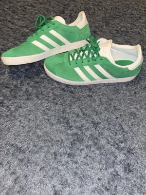 Gröna Adidas Gazelle sneakers - Säljer ett par gröna Adidas Gazelle sneakers i mocka med vita detaljer och klassiska tre ränder på sidan. Skorna har vit sula och hälkappa med Adidas-logga. Lågt skaft och snygg retrostil som passar till det mesta.