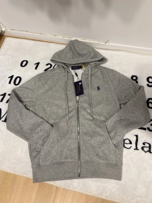 Grå hoodie från Polo Ralph Lauren - Snygg grå zip-hoodie från Polo Ralph Lauren med dragsko i huvan och klassisk blå/lila logga broderad på bröstet. Tillverkad i mjuk bomullsblandning och har två stora fickor framtill. Perfekt för en chill och stilren look.