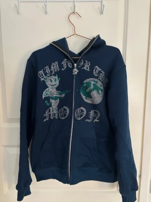 Mörkblå hoodie med tryck och dragkedja - Mörkblå hoodie med dragkedja och huva. Framsidan har ett stort tryck med texten 'Aim for the moon', en tecknad figur och en jordglob i grönt och vitt. Perfekt för en avslappnad och cool stil.