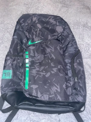 Svart och grönNikeelite ryggsäck med mönster - Säljer en svart ryggsäck från Nike med grått abstrakt mönster och gröna detaljer, inklusive Nike-loggan och ränder på framsidan. Ryggsäcken har flera fack med dragkedja och en förstärkt botten i konstläder. Perfekt för skola eller träning.