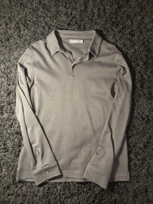 Grå långärmad tröja Dressmann - Snygg grå långärmad tröja från Dressmann med diskret fiskbensmönster. Tröja har klassisk krage, knappar vid ärmslut och är tillverkad i ett mjukt tyg som känns bekvämt mot huden. Perfekt för en stilren look.