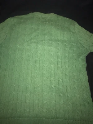 Grön kabelstickad tröja Polo Ralph Lauren - Snygg grön kabelstickad tröja från Polo Ralph Lauren med rund halsringning och ribbade muddar. Tröjan har ett diskret blått logobroderi på bröstet och är tillverkad i mjukt material. Perfekt för dig som gillar klassisk stil.