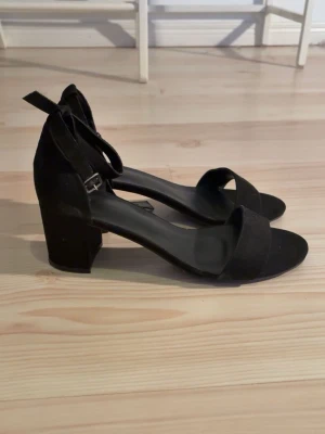 Svarta sandaler med blockklack - Stilrena svarta sandaler med bred rem över tårna och justerbart ankelband. Skorna har en stabil blockklack och öppen tå, vilket ger en trendig och enkel look. Materialet ser ut att vara mockaimitation och innersulan är svart. Perfekta för dig som gillar minimalistisk stil.