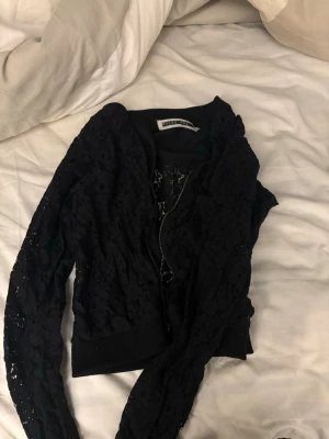 Svart spetskofta från Ivyrevel - Snygg svart kofta från Ivyrevel med dragkedja framtill och genomgående spetsdetaljer. Långärmad modell med ribbade muddar vid ärmslut och nederkant. Perfekt för att lyfta din outfit med en trendig touch.