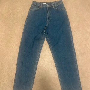 Monki Taiki blå high waist jeans - Säljer ett par blå Monki Taiki jeans med high waist och balloon leg. Jeansen är gjorda av ekologisk och återvunnen bomull. Klassisk femficksmodell med snygg passform och läderpatch bak i midjan. Perfekta för en trendig och avslappnad stil.