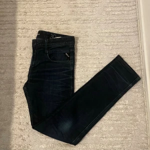 Mörkblå jeans från Replay  - Säljer ett par mörkblå jeans från replay med smal passform och klassisk femficksdesign. Jeansen har subtila slitningar och är tillverkade i ett stretchigt denimtyg för extra komfort. Perfekta för en avslappnad och stilren look.