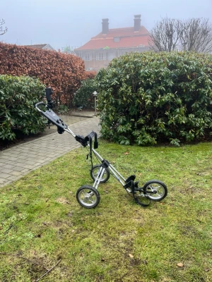 Trehjulig golfvagn i metall - Säljer en smidig och lätt trehjulig golfvagn i silverfärgad metall med svarta detaljer. Vagnen har justerbart handtag, hållare för scorekort och mugghållare. Perfekt för att enkelt transportera golfbagen runt banan.