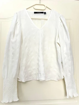 Vit blus med v-ringning vero moda - Stilren vit blus från vero moda med v-ringning och långa puffärmar. Ärmsluten har bred smock och lätt volangkant. Blusen är tillverkad i ett lätt strukturerat tyg som ger en elegant känsla. Perfekt att matcha med jeans eller kjol för en trendig look. Använda 1 gång!! 