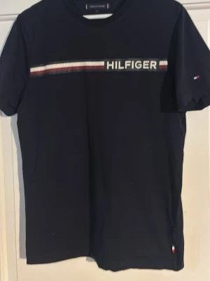 Svart t-shirt från Tommy Hilfiger - Snygg svart t-shirt från Tommy Hilfiger med klassiskt logoband i vitt, rött och blått över bröstet. T-shirten har rund hals och korta ärmar, perfekt för en avslappnad och stilren look. Materialet är mjuk bomull som känns skönt mot huden.