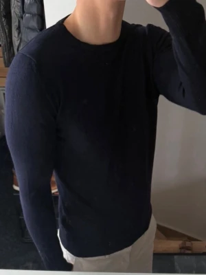 Mörkblå tröja från J.Lindeberg - Snygg mörkblå tröja från J.Lindeberg med rund halsringning och långa ärmar. Tröjan är tunnstickad och har ribbade muddar vid ärmslut och nederkant. Perfekt för en stilren och enkel look. 100% merinoull. Storlek m men har krympt så sitter som xs