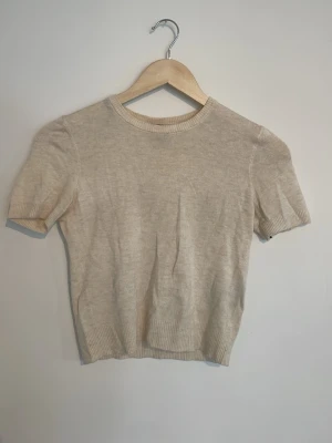 Beige stickad kortärmad topp - En stilren beige stickad topp med korta ärmar och rund halsringning. Toppen har ribbade kanter vid ärmslut, hals och nederkant. Perfekt för en enkel och clean look.❣️