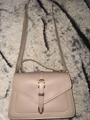 Beige axelväska  - Snygg beige axelväska i slätt konstläder med justerbart axelband och dekorativ guldfärgad spänndetalj på locket. Väskan har en stilren, rektangulär form och passar perfekt till en trendig vardagslook.