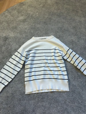 Vit stickad tröja med svarta ränder - Vit stickad tröja från 157 med svarta horisontella ränder över hela tröjan. Tröjan har rund halsringning och långa ärmar. Perfekt för en avslappnad och stilren look.