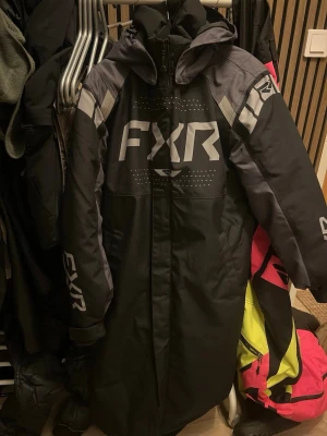 Svart depårock med huva från FXR - Säljer en svart depårock från FXR med grå detaljer och stora logotexter på bröstet och ärmarna. Jackan har huva, dragkedja framtill och reflekterande detaljer på ryggen. Perfekt för dig som vill ha en väldigt varm jacka på skoterturen eller över din skoteroverall. Är xxs men passar även M/L. Herrmodell