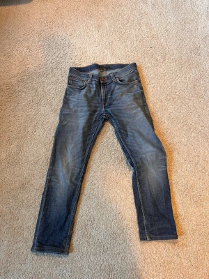 Blå jeans från Nudie Jeans - Säljer ett par klassiska blå jeans från Nudie Jeans med raka ben och fem fickor. Jeansen har snygga slitningar och kontrastsömmar samt Nudie-logga på bakfickan. Tillverkade i bomull för en skön känsla och perfekt passform.