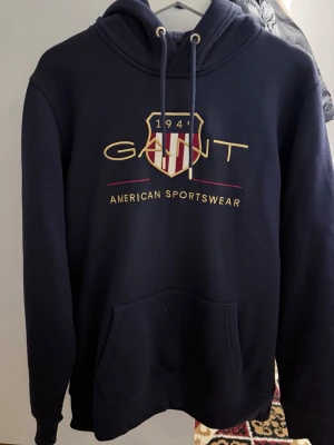 Marinblå hoodie från GANT - Snygg marinblå hoodie från GANT med broderad logga och texten 'American Sportswear' på bröstet. Klassisk huva med dragsko och stor magficka framtill. Tillverkad i mjuk bomullsblandning, perfekt för en avslappnad och sportig stil.